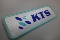 白地に青い“KTS”ロゴと楕円形マークが加工された水色の糸で縁取りされた長方形オリジナルプリントワッペンの制作事例。