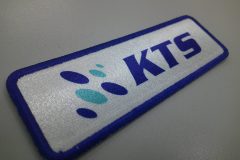 白地に青い“KTS”ロゴと楕円形マークが加工され青糸で縁取られた長方形オリジナルプリントワッペンの制作事例。