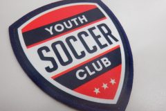赤・白・紺の配色で“YOUTH SOCCER CLUB”の文字が入った盾形のオリジナルプリントワッペンの制作事例。下部に3つの白い星がある。