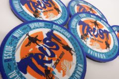 中央に“TRUST”の文字と握手のモチーフ、ロープで下降する人物のシルエットが描かれた青い縁取りの円形オリジナルプリントワッペンの制作見本を複数枚並べた画像。