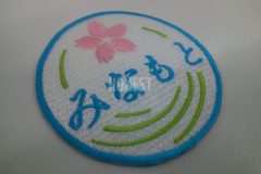 白のクロス生地に水色でみなもとの文字と桜と葉をイメージした線をピンク、黄緑の糸で表現したオリジナル刺繡ワッペンの制作事例。