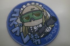青地に水色の縁取りの円形ワッペン。中央に金髪のキャラクターがメイド服を着て緑のゴーグルをかけ、黒い銃を構える姿が描かれたオリジナル刺繡ワッペンの制作見本。