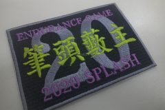 黒地に灰色の縁取りがある長方形刺繍ワッペン。中央に大きな灰色の数字『20』が配置され、その上下に紫文字と黄緑色の漢字が重なるように加工されているオリジナル刺繡ワッペンの制作事例。