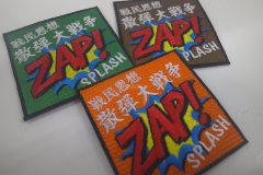 3枚の四角形のワッペンが並んでいる。いずれも同じデザインで中央に大きく「ZAP！」と前面に飛び出すような表現で描かれたオリジナル刺繡ワッペンの制作事例。緑生地・茶色生地・オレンジ生地の3色展開。