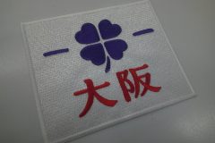 白のエンブクロス生地に紫の糸でクローバーを加工。その下に赤糸で大阪の文字を刺繍したオリジナルワッペンの制作事例。