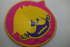 吹き出しマークの中に眉間に皺の寄った男の子のキャラクターを刺繡したオリジナル刺繡ワッペンの制作事例。ピンク色の生地に黄・紫で刺繍。
