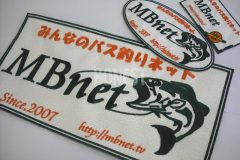 アイボリーカラーのナイロンエンブクロス生地にモスグリーンとオレンジの糸でバス釣りチームのロゴを刺繍した、オリジナルワッペンの作品事例。