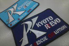 ロゴマークを大きく刺繍し、団体名を白糸と赤糸でアクセントを付けて刺繡した、オリジナル刺繡ワッペン2種を並べた実例。