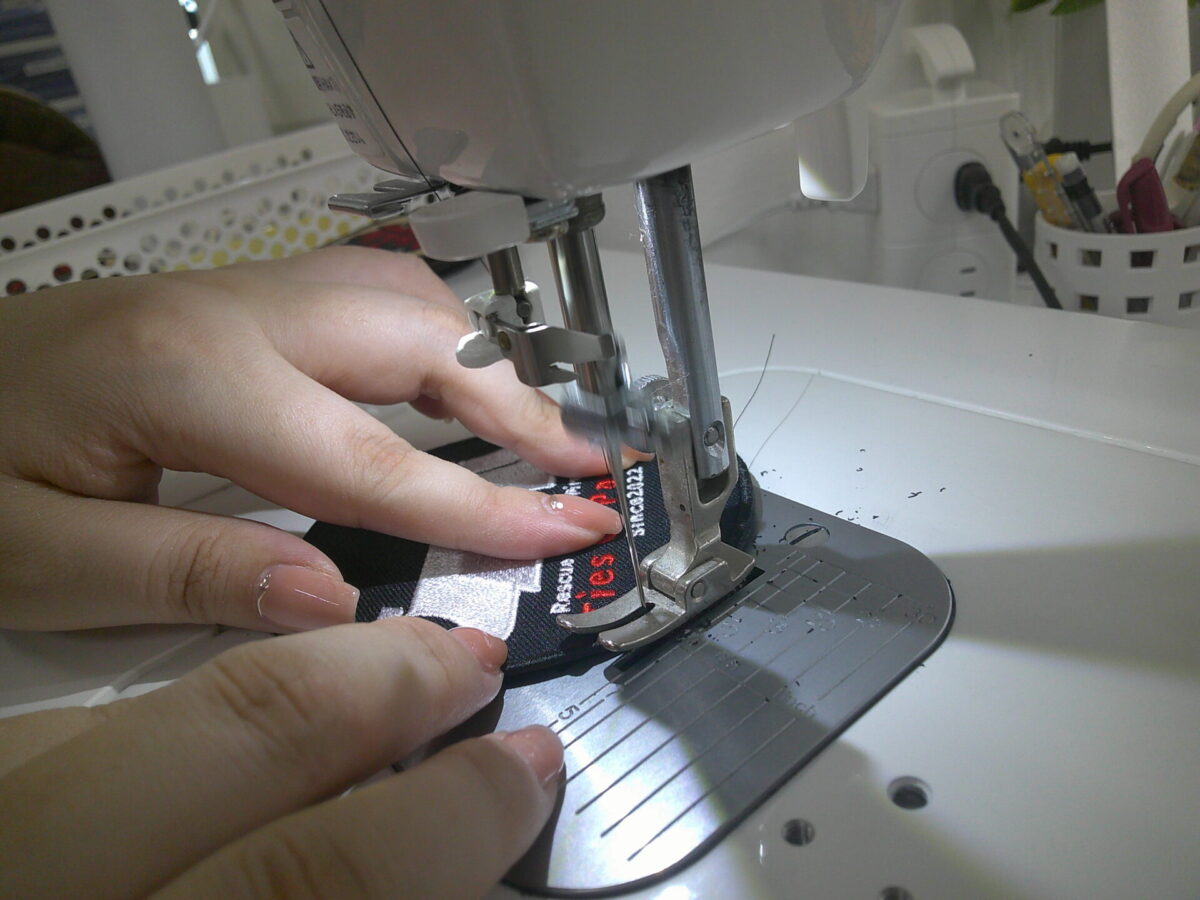 円形のワッペンに工業用ミシンでベルクロ（黒）を縫い付けている作業工程。