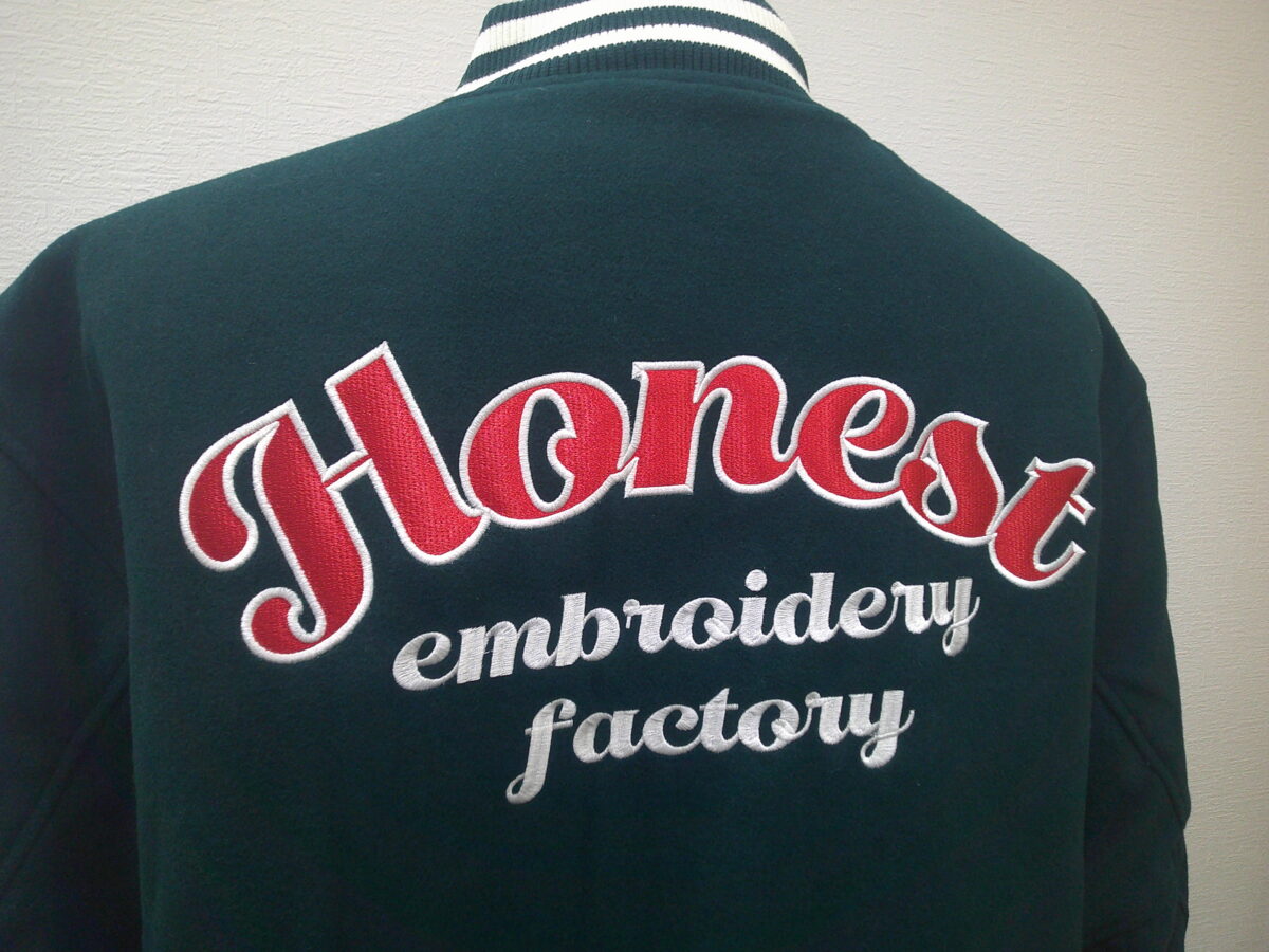 メルトンジャケット背面に赤生地をベースとした縁取り刺繍。その下に筆記体で白の文字刺繍を加工した制作実績。