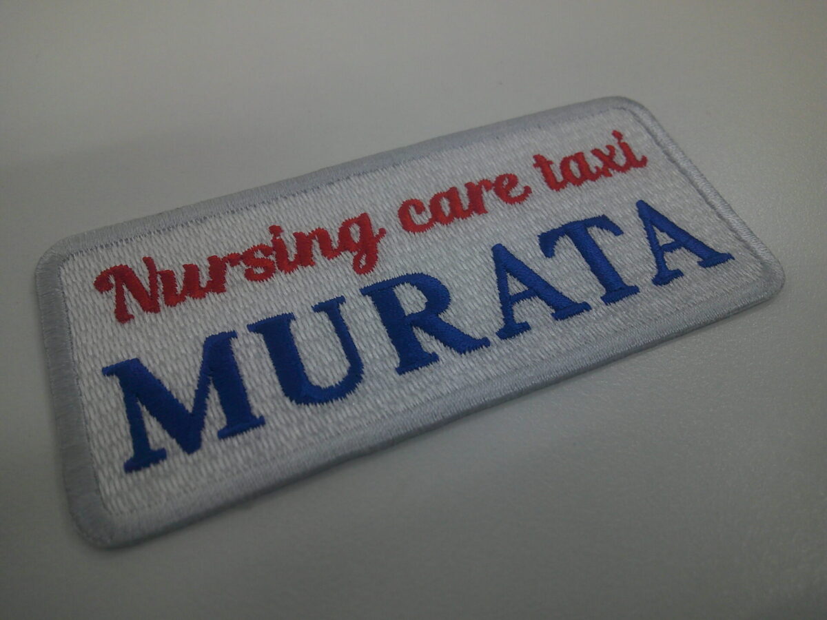 白生地に赤と青とグレーの糸で刺繍をした、介護タクシー会社の文字ワッペン。
