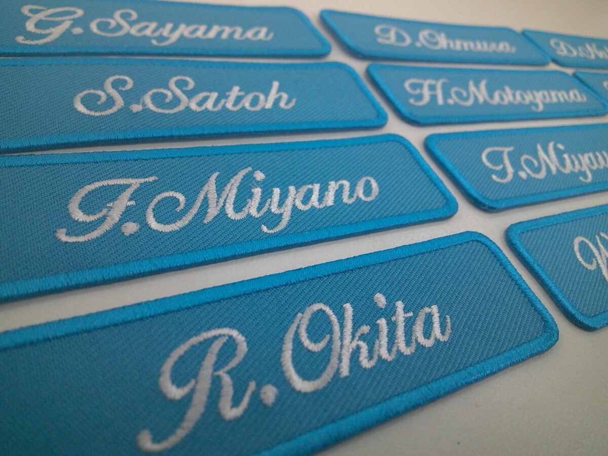 それぞれ違う個人名が白糸で刺繍された、文字ワッペン。ターコイズ生地に白糸の刺繍。
