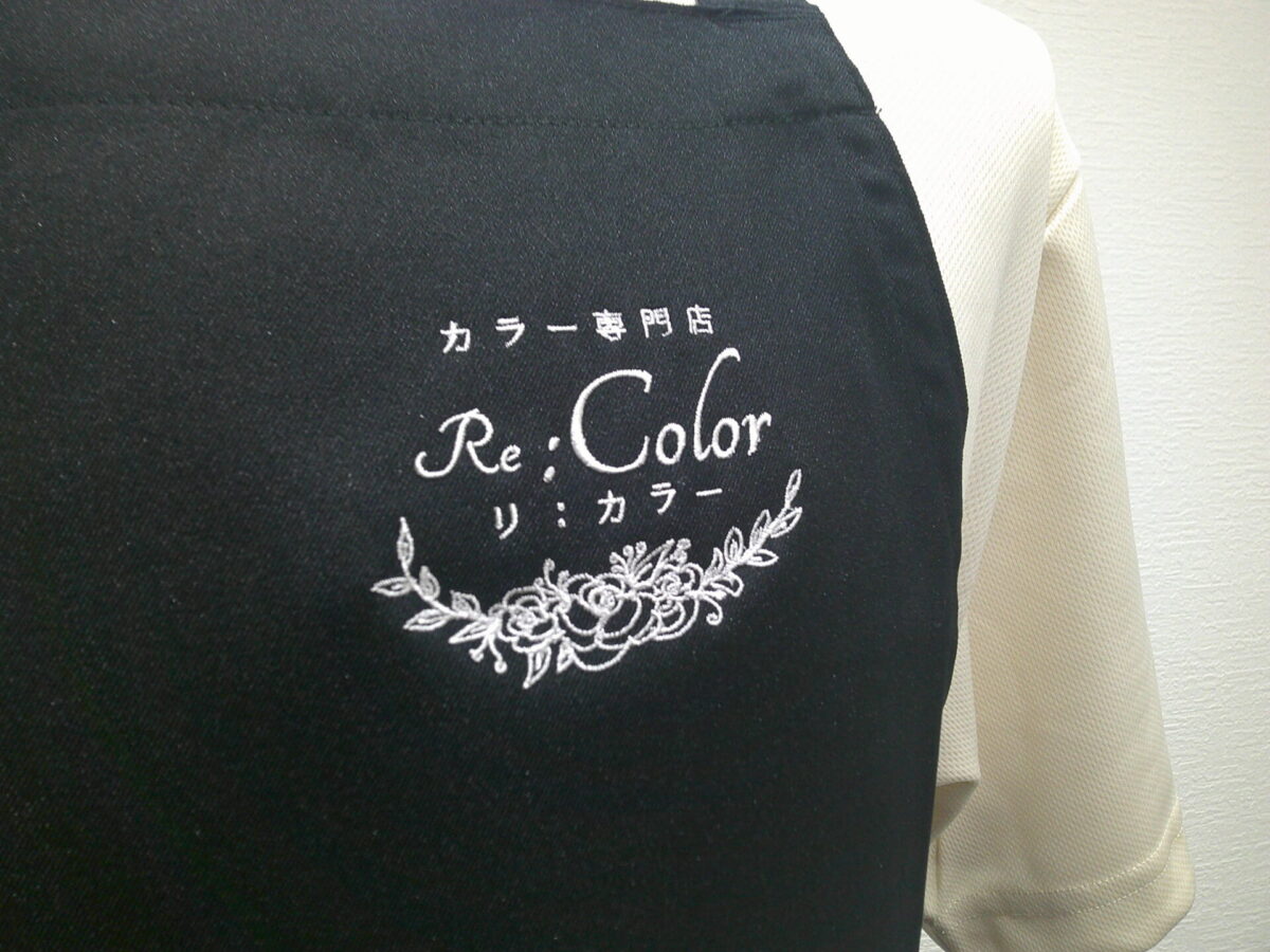 エプロン(FK7168)の左胸に白糸で美容室のロゴ刺繍(フローラルデザイン)を加工した制作事例|左上から見たアップ