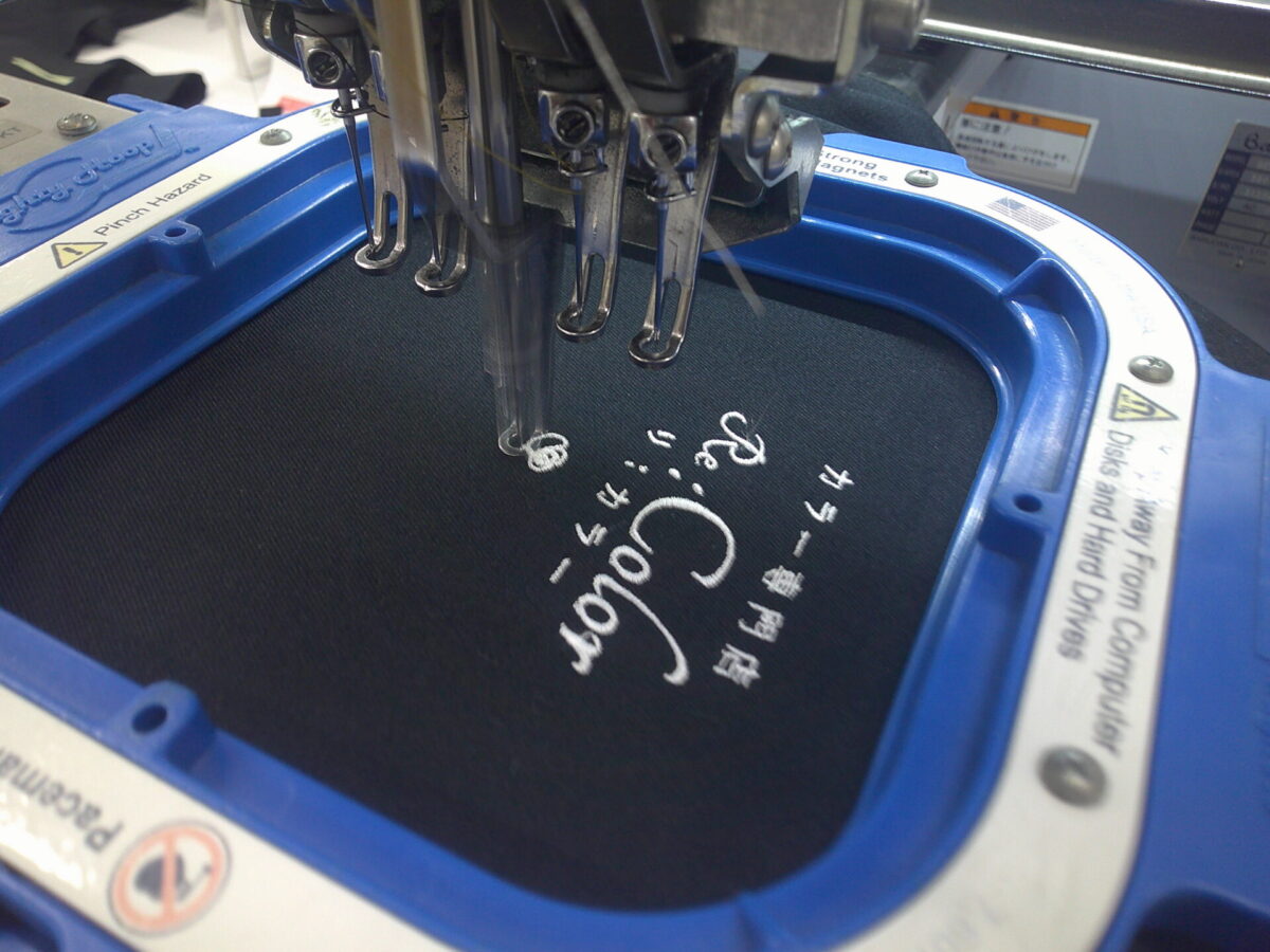 黒のエプロンの左胸に美容院のロゴデザインを白糸単色で刺繍加工をしている、作業工程。