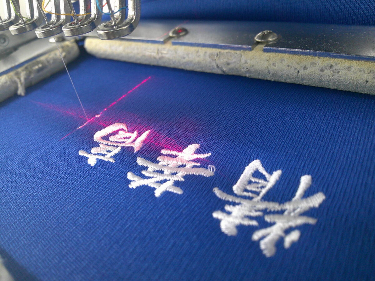 ブルーのドライポロシャツの左胸にクランプ刺繍機にて、学校名を刺繍している作業工程。刺繍糸はホワイトを使用。