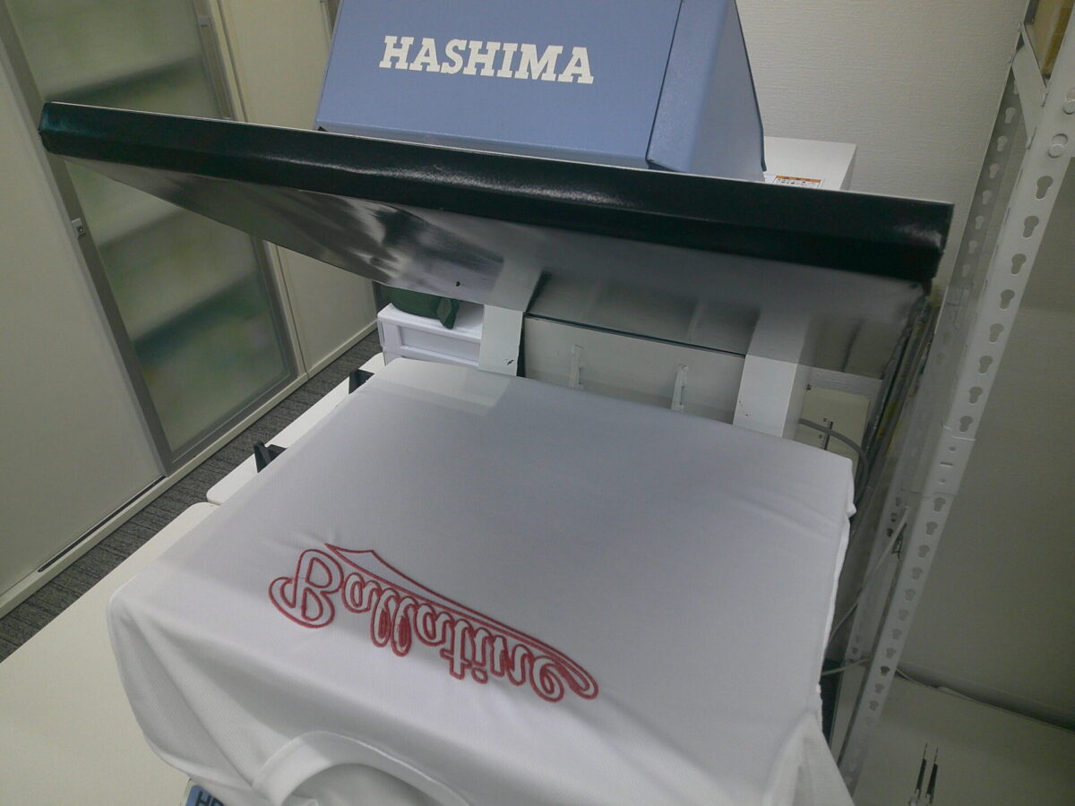 縁取り刺繍加工後、圧着機で熱プレス仕上げを行っている作業工程。