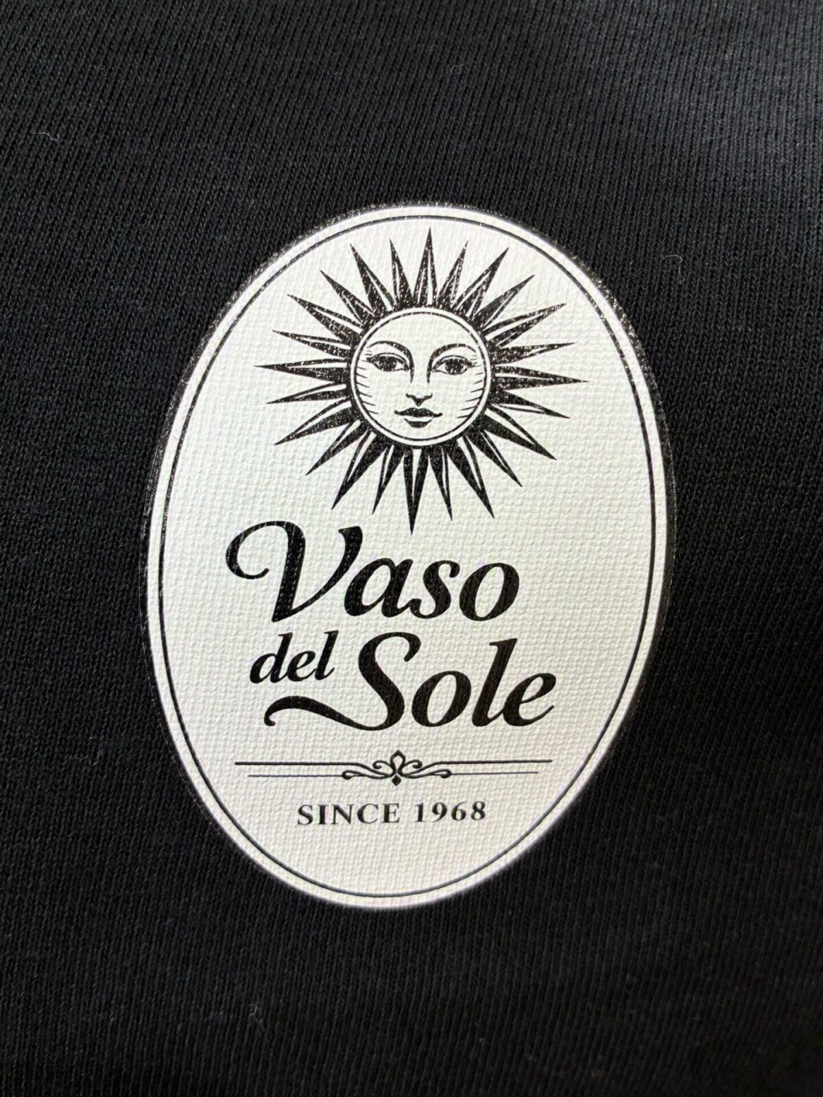 黒のTシャツにイタリア料理店のロゴを白ベースにてエンブレムプリント加工した制作事例。真上からのアップ。
